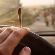 Une main tient un joint de cannabis et le volant d'une voiture. 