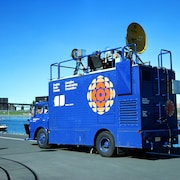 Près du bassin olympique, car de reportage de Radio-Canada avec le logo de la pizza multicolore. Des techniciens sont assis sur le toit du véhicule.
