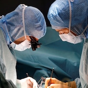 Dans une salle d'opération, deux personnes portant des combinaisons bleues et des masques chirurgicaux sont face à face, et sont penchées au-dessus d'une table d'opération où est couché un patient, hors du cadre de la photo.