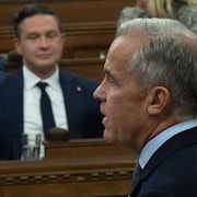 Mark Carney debout lors de la période des questions avec Pierre Poilievre en arrière-plan.