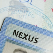 Le coin d'une carte bleue où apparaît le mot « Nexus » posé sur une page d'un passeport canadien décoré d'un tampon du département américain de la sécurité intérieure qui autorise l'entrée d'un voyageur.