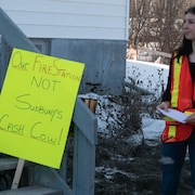 Une femme porte un gilet rouge et à sa droite, il y a une affiche qui réclame le maintien de la caserne de pompiers de Skead