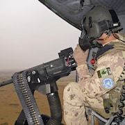 Un soldat canadien est assis dans un hélicoptère, devant un fusil mitrailleur. 