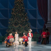 Des enfants portant le costume de la police montée gambadent sur une scène devant des danseurs de ballet et un immense sapin de Noël.