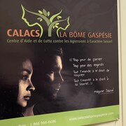 L'affiche de l'organisme à l'entrée du bureau. La porte est ouverte et on peut voir un fauteuil et une plante