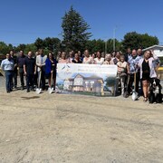 Une vingtaine de personnes réunies pour la première pelleté de terre. Ils se tiennent derrière une affiche du projet. 