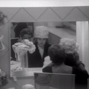 Une femme essaie un chapeau devant un miroir et une vendeuse ajuste son chapeau derrière elle. 