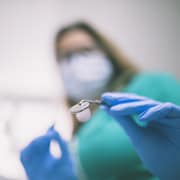 Une femme qui porte un masque de papier devant sa bouche et des gants de plastique tient dans ses mains des outils pour les soins dentaires. 
