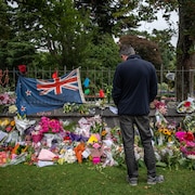 Un homme se recueille devant des fleurs et un drapeau de la Nouvelle-Zélande, déposés sur le mur d'un parc.