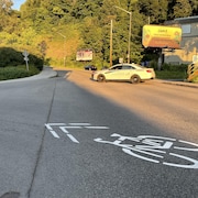 Une voiture de police est stationnée au milieu d'une rue asphaltée, bloquant la circulation, à l'intersection d'une route. Un pictogramme de vélo est peint au sol. Le soleil rasant éclaire la scène, projetant de longues ombres. 