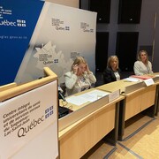 Trois personnes sont assises derrière des bureaux en demi-cercle.