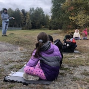 Des élèves écoutent une enseignante, assis sur le terrain de l'école.