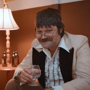 L'humoriste Maxime Gervais dans son personnage de Claude Crest, assis dans un salon à l'esthétique des années 1950, verre à la main. 