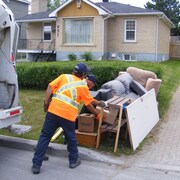 Deux employés ramassent de gros déchets sur le bord de la rue.