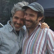Deux hommes s'enlacent derrière une scène de festival extérieure. 