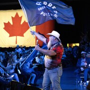 Un homme barbu coiffé d'un chapeau tient un drapeau en signe de soutien à Pierre Poilievre tandis qu'une assemblée de délégués conservateurs réagit à un discours.