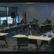 Treize personnes sont assises autour d'une table de réunion, devant un écran géant.
