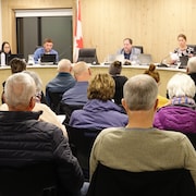 Des gens assis, de dos, font face à la table du conseil de ville où siègent les élus.