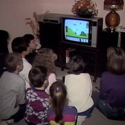 Un groupe d'enfants sont tournés vers un écran de téléviseur sur lequel se déroule une partie du jeu Mario Bros sur console Nintendo.