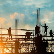 Les silhouettes de personnes sur une partie surélevée d'un chantier de construction sont vues lors d'un coucher ou d'un lever de soleil. Ces personnes portent des casques.