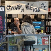 Un homme derri&egrave;re un kiosque o&ugrave; il est inscrit &laquo; Consultez un a&icirc;n&eacute; pour 25 cents &raquo; &eacute;coute attentivement une femme vue de dos qui est assise sur un tabouret devant lui.