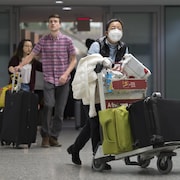 Des voyageurs avec leurs bagages dans un aéroport. Une voyageuse porte un masque.