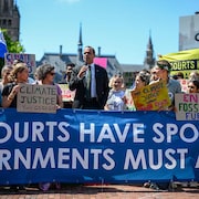 Un homme parle dans un micro, entouré d'une foule de gens qui tiennent des pancartes et des banderoles pour réclamer davantage de justice climatique.