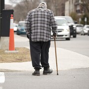 Un homme âgé marche sur le trottoir avec une canne, dans une rue où passent des voitures.