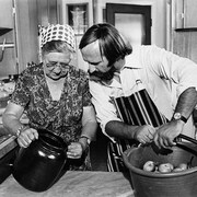 Dans une cuisine, Madame Ernest Dubé, pomicultrice de Rougemont, et l'animateur Jacques Houde sont devant une planche de travail sur laquelle sont posés une cruche et un panier de pommes.