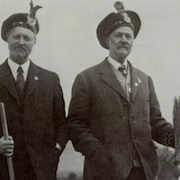 Deux hommes portant le costume traditionnel écossais et tenant chacun un balai. 