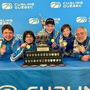 Quatre jeunes joueurs de curling et leur entraîneur exhibent fièrement leur médaille d'or et leur trophée.