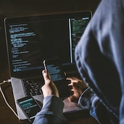 Un pirate informatique assis en face d'un ordinateur et tenant un téléphone portable.