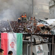 Des hommes avec des dossards orange éteignent un incendie d'un bâtiment où il y a un géant drapeau de l'Iran.