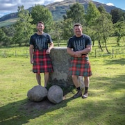 Deux hommes en kilt posent avec de grosses pierres rondes à leurs pieds.