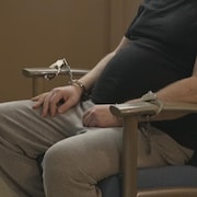 Un homme portant un chandail noir et un pantalon gris est menotté alors qu'il est assis dans une chaise. 