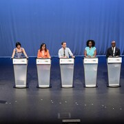Les six candidats sur scène, derrière des lutrins. À gauche, le modérateur Steve Paikin les regarde.