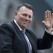 Dennis King sourit et fait un salut de la main gauche.