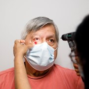 Un homme âgé regarde droit devant lui pendant que la main aux ongles vernis de son médecin abaisse sa paupière droite pour examiner son oeil.