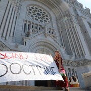 Deux personnes tiennent une large banderole où il est inscrit « Rescind the doctrine » devant la basilique Sainte-Anne-de-Beaupré.