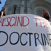 Deux personnes autochtones tiennent une banderole où il est écrit « rescind the doctrine ».