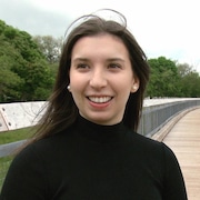 Une jeune femme debout sur une passerelle de bois regarde à droite et sourit, le vent dans ses longs cheveux foncés.