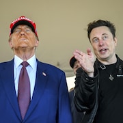 Donald Trump, portant une casquette rouge MAGA, regarde au loin alors qu'Elon Musk lui parle en gesticulant.