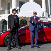 Debout devant une voiture sport Tesla rouge stationnée devant la Maison-Blanche, Donald Trump fait un geste avec ses mains. Elon Musk est debout à sa droite et le regarde.