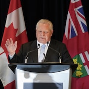 Doug Ford, levant la main, debout derrière un micro, entouré des drapeaux du Canada et de l'Ontario.