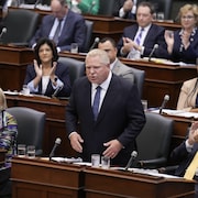 Le premier ministre se lève et répond en Chambre devant ses collègues qui applaudissent.
