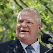 Gros plan sur le premier ministre Ford, dehors, entouré de verdure, devant un micro.