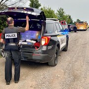 Un policier est debout à l'arrière d'un véhicule de police dont le hayon est ouvert. Plusieurs autres véhicules d'urgence sont stationnés sur le côté d'une route en gravier.
