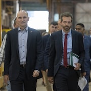 Jean-Yves Duclos et Joël Lightbound en compagnie du maire de Lévis, Gilles Lehouillier. Tous les trois marchent avec des hommes en complet derrière eux. Ils sont dans une usine. 