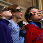 Quatre enfants avec des lunettes de protection orange et noire en carton. Leurs têtes sont levées vers les cieux.