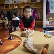 Un enfant les avant-bras sur une table dans une classe pose pour la photo en tenant une collation.